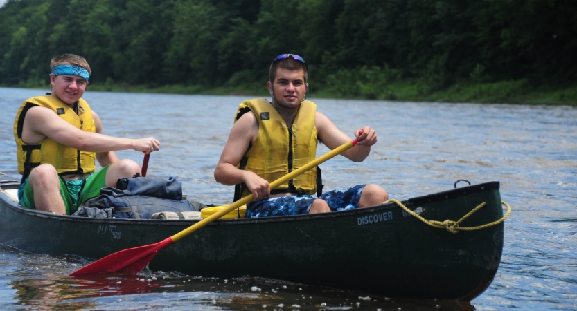 Two people wearing yellow life jackets paddle a canoe on calm water